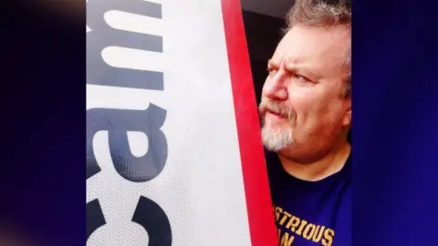 Steve Jollands with a short grey beard and moustache holding up the RAF Scampton sign and looking at it whilst frowning