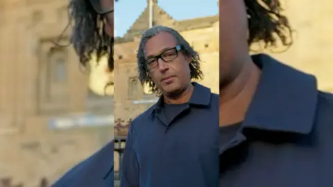 A man, Professor David Olusoga, with long dark hair, glasses and wearing a blue top.