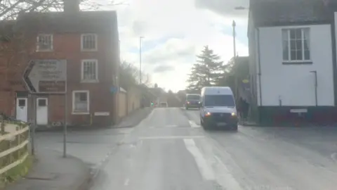 A road travels straight and over the brow of a hill. There is a junction where you can turn both left and right. Either side of the road are houses, and there is a road sign on the left hand side. There is a white van driving on the road.