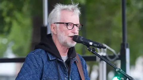 Billy Bragg sings into a microphone as he performs on an outdoor stage. He is wearing a denim-coloured jacked and black-rimmed glasses. He has messy grey hair and a short, full beard.