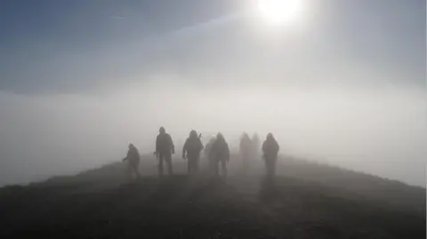 BBC Weather Watchers/StorytellerK Walkers on the hills are seen in mist and fog and some sunshine on Saturday. The group look to be wearing outdoor gear and are seen in silhouette against the sun.