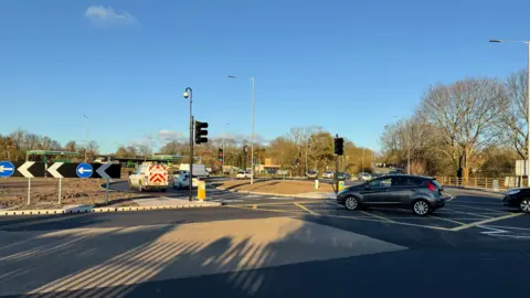 BBC The Dunball roundabout with cars going around it. The sun is shining on the tarmac.
