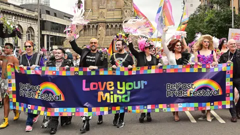 Pacemaker Organisers of Belfast Pride, and Lord Mayor Micky Murray, hold a banner with the organisation's banner on it with 'Love Your Mind' written in rainbow font. Many of the parade's participants are holding rainbow and progress flags, alongside the asexual and transgender flag.