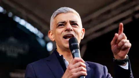 Mayor of London Sadiq Khan speaks on stage. He is wearing a navy jacket and an open-necked white shirt. 