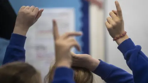Children's raised hands against the blurred backdrop of a school classroom.