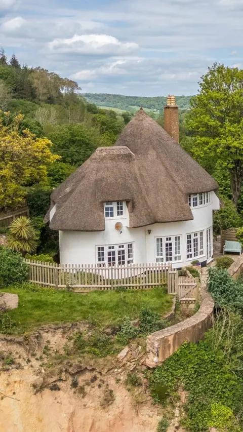 A picture looking from above down at the property and the cliff top. It is a large White House, with a brown roof. There is grassland and trees around the property.