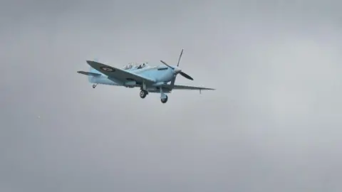 Ian Knight A blue Spitfire in flight, against a grey sky.