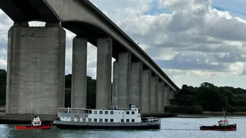 Julien Jourdain A large white boat is towed by a smaller boat underneath a large bridge on a river.