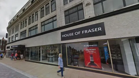 Google House of Fraser in Darlington. It is a grey three-storey building. There are make-up brands in the shop front. A woman wearing a blue shirt and jeans in passing by. A couple of people are entering the store. 