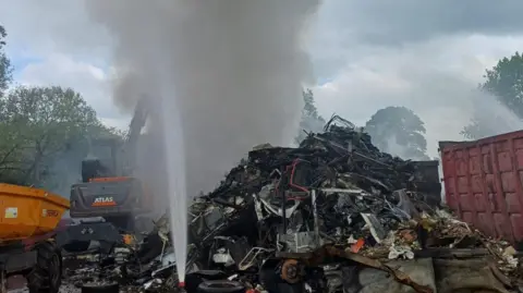 London Fire Brigade A close up of the fire at the metal recycling centre. Thick, grey smoke is rising from the pile of metal.
