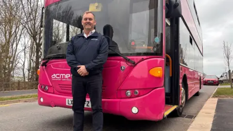 James Palmer stands in front of his parked bus. He is wearing his uniform, a zip-up top and dark trousers.