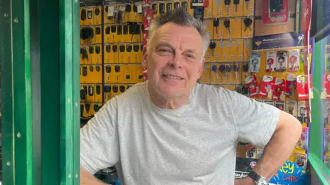 Jonny Humphries/BBC Gary Hartles, who has grey hair and is wearing a light grey t-shirt, smiles at the camera from behind the counter of a shop. A number of keys are on a yellow rack behind him. 