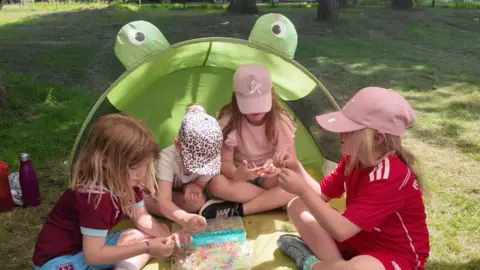 Three schoolgirls sitting outside under a shaded area, shaped like a frog. They appear to be playing a game. There are water bottles nearby.