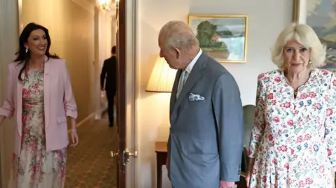 PA Media A man in a grey suit and woman in floral-patterned dress are greeted by another woman in a different floral-patterned dress and a pink jacket