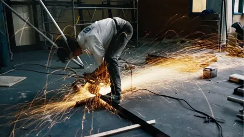 Kendal Wall A man crouches over metal framework, which he appears to be cutting with a rotary metal saw. Sparks are spraying out from the point of contact, forwards and backwards. He is wearing dark grey jeans and a white, long-sleeved t-shirt. Beyond him is a steel scaffolding frame.