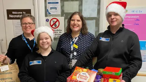 Our 24 Special Advent Days A woman in a black jacket holds a paper bag in a hospital ward. She stands next to a woman and a man wearing Santa hats. The man is holding a open hamper of biscuits, tea and snacks. A woman in a navy dress stands between them smiling at the camera.