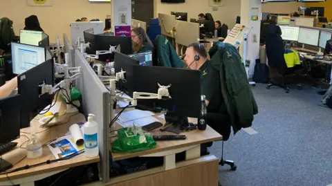 NIAS control room. People in ambulance uniforms are sitting at desks in front of computer screens wearing headsets.