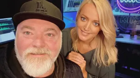 A middle aged man with a black baseball cap and grey beard and a blonde-haired younger woman smile at the camera