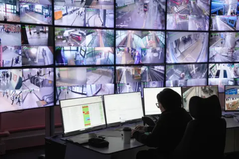 BBC/Andy Alcroft A large number of TV screens show live CCTV footage. A woman is sat at a desk watching it. 