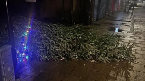 A Christmas tree lying on its side on the street, with only a few lights still working. The street is dark, with scattered puddles. 