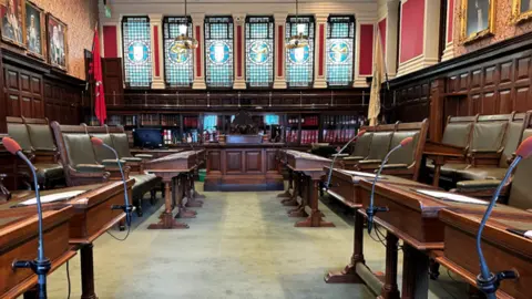 BBC The inside of the House of Keys Chamber, which has green leather clad wooden chairs and benches on either side and a bookcase and stained glass panels behind the speaker's desk.