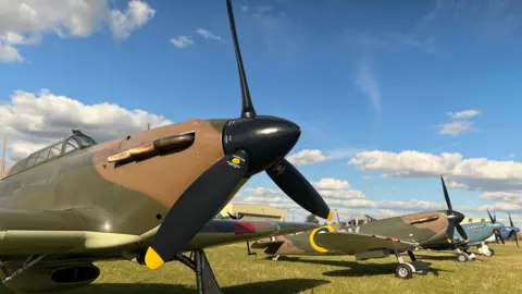 Emma Baugh/BBC Several warbird planes in a line, they are painted in dark green and brown camouflage with black propellers. They are positioned on grass and behind them is a bight clue sky with some white clouds. 