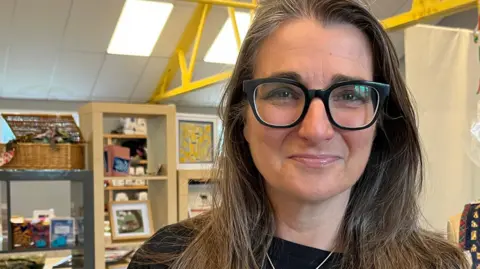 A head and shoulders image of Melissa Santiago-Val. She is wearing a black top and a necklace and glasses. She is looking directly into the camera. Behind her is some shelving with various items on it and to her left is a headless mannequin with handmade jewellery and clothing hanging on it.