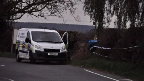 NEMM Police van, parked by a rural road, with trees and a high hedge, with police cordon tape visible along the hedge