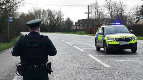A police man, with his back to the camera, stood on a road with a police car beside him. There are rows of houses and greenery in background.