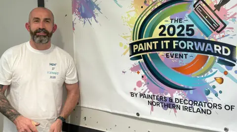 Paintworks NI Gerard Donaghy is standing in front of a white wall, wearing a white t-shirt and is smiling at the camera. He has a bald head and a dark beard with grey hair through it.