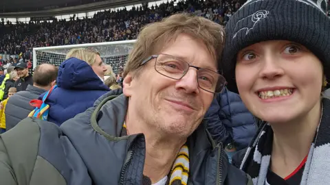 SUPPLIED Man with glasses and puffer jacket sitting next to teenage child in bobble hait and a football scarf in a football stadium during a football match