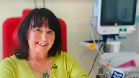 Carol Robson is getting her bloods taken as part of the trial. She is smiling while seated with her sleeve rolled up and surrounded by medical equipment. She has dark shoulder-length hair and wears a lime green v-neck top and a chain with a large rectangular green stone.