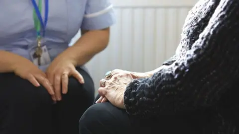 Getty Images A nurse in a blue uniform sits beside a woman in her home