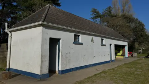 The toilet block is white with a grey roof. There is a blue strip at the bottom of the building.