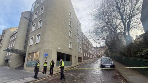 Iona Young The street is on a steep slope. There is yellow police tape at the bottom. There are four police officers in high vis guarding the site.