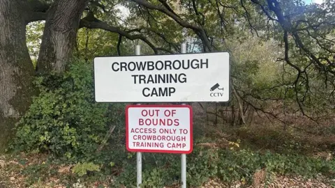 A sign reading Crowborough Training Camp, above a second sign reading: Out of Bounds, access only to Crowborough Training Camp. There is woodland beyond the signs