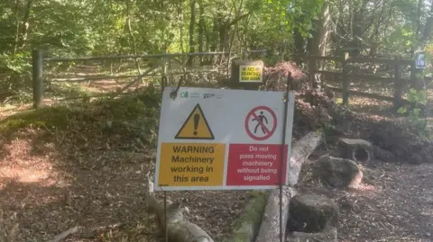 DUNCAN HILL/BBC The closed off entrance to the bike park warning visitors about ongoing machinery work. The scene is located in a woodland environment with tall mature trees and dense green foliage.