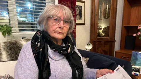 A 79 year old woman sat on her sofa in her living room. Lady has a grey short bob and full fringe and is wearing glasses looking into the lens and holds the letter from the council in her left hand. 