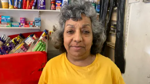 Jade Taylorson/BBC Jyoti Broadhurst, in her 60s, with greying black hair and a yellow t-shirt, smiles into the camera in a convenience shop. 