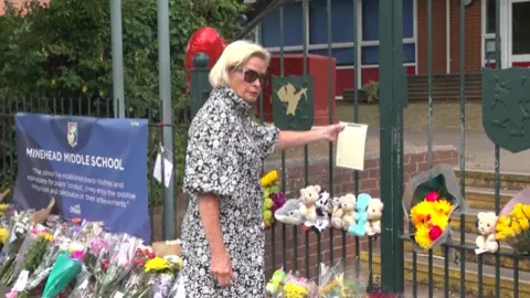 PA Media Rachel Gilmour is wearing a black and white dress. She is standing by the school gates which are covered in flowers and toys. She is putting a note on the gate.