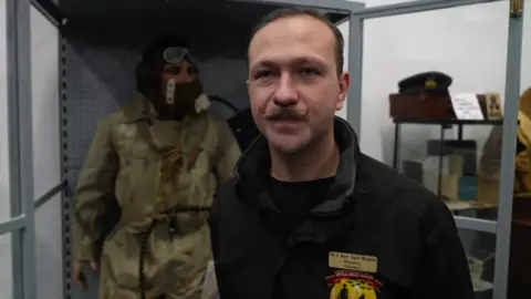 A man stands in front of a museum display, including a mannequin wearing an RAF pilot's uniform. He has short brown hair and a moustache that curls up at the ends. He is wearing a black t-shirt, a black collared fleece and a gold name badge. 