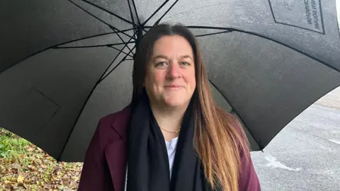 Kimberley Whitehead is pictured wearing a purple blazer and navy scarf. She is standing next to a road on a rainy day and is underneath a large black umbrella.