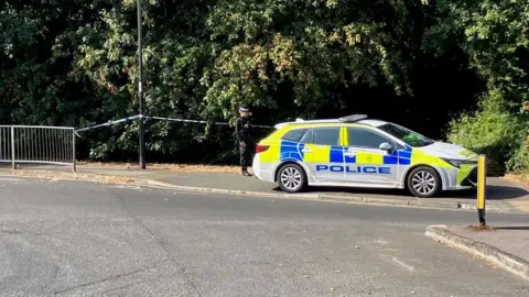 Police car and tape across a wooded area off a roundabout with a sign to Swaythling.