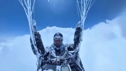 Douyin/CCTV A paraglider covered in ice, wearing a helmet, goggles, big gloves and equipment, holding onto the ropes, floats in front of a blue sky and white clouds