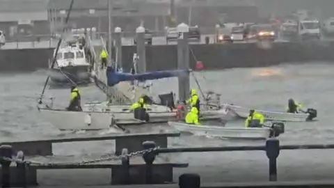 Samantha Jane Boats in Victoria Marina rescuing a large sailing boat. There are three boats surrounding the vessel with people wearing hi-vis. 