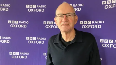 A man is standing in front of a BBC Radio Oxford background. He is wearing glasses and a black shirt.