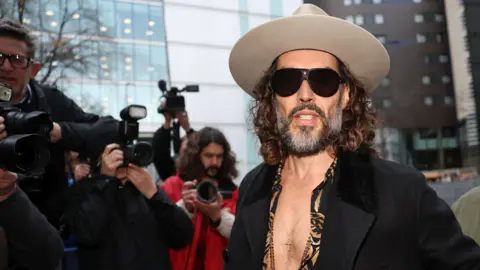 Reuters Russell Brand arriving at Southwark Crown Court wearing a light grey fedora hat, dark glasses, a leopard print shirt unbuttoned halfway down his chest, a cross on a necklace, and a dark jacket