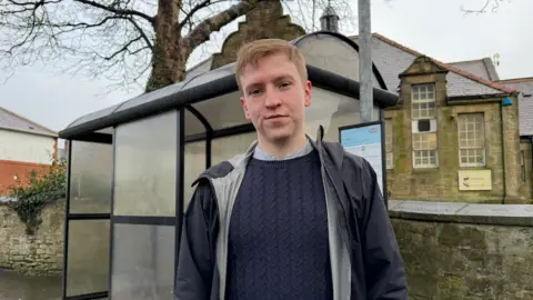 Councillor Declan Mulholland. He has fair blond hair and wears a navy blue jumper and a black and grey jacket. He is standing in front of a bus stop in a village.