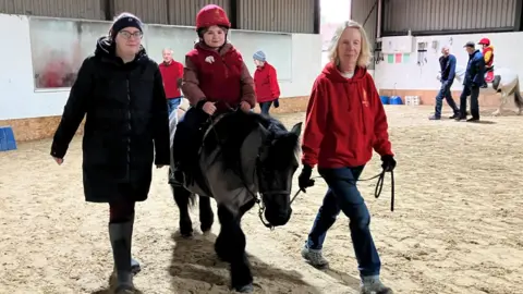 A service user and their mum on one of the ponies at the East Park Riding for the Disabled Group in Lingfield, Surrey. The black pony is being led by one of the volunteers at the East Park Riding for the Disabled Group in Lingfield, Surrey. The volunteer is wearing a red hoodie, while the mum is wearing a black puffer coat and glasses.