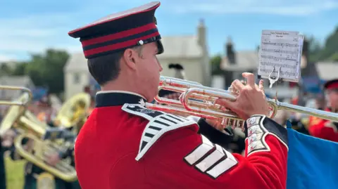 BBC A member of the Band of the Princess of Wales's Royal Regiment following the sheet music of the of the Manx National Anthem. He is wearing an embellished red jacket and a black and red peaked cap.